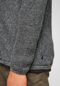 Pull gris texturé avec un motif tricoté, présentant une étiquette logo noire à l'ourlet et des poignets côtelés. La couche intérieure est sombre.