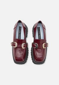 Burgundy patent leather loafers with gold-tone buckles and thick black rubber soles, featuring a sleek shape and smooth texture.
