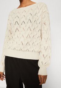 Krämfärgad stickad tröja med en strukturerad chevronmönster, bred halsringning och ribbad mudd och nederkant. Kombineras med svarta byxor.