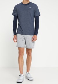 Mężczyzna ubrany w granatową koszulę z długim rękawem pod niebieską koszulą z krótkim rękawem, szare szorty i białe sneakersy, stojący na białym tle.