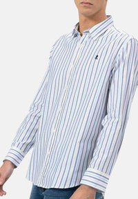 Camisa de botones a rayas con líneas verticales en azul claro, lavanda y blanco. Incluye un pequeño logo en el bolsillo del pecho.