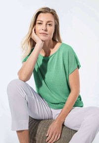 Grünes Kurzarm-T-Shirt aus weichem Stoff, mit entspanntem Schnitt und Rundhalsausschnitt. Kombiniert mit hellgrauten Hosen, sitzt auf einer strukturierten Oberfläche.