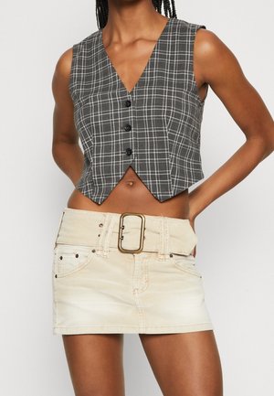 Gilet sans manches à carreaux noir et blanc avec boutons, associé à une mini-jupe beige dotée d'une large ceinture et d'une grosse boucle.