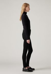 Moteris su ilgais šviesiais plaukais, vilkinti juodą golfą, juodas aptemptas kelnes ir juodus loafers, stovi profiliu prieš pilką foną.