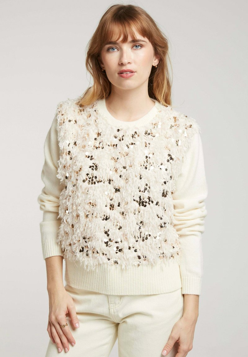 Pull léger beige en tissu texturé et duveteux, avec un motif superposé de paillettes. Les poignets et l'ourlet côtelés ajoutent de la définition.