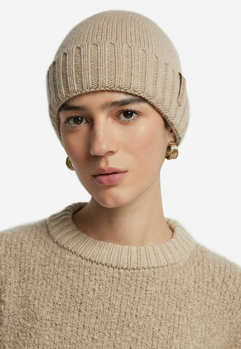 Beige stickad mössa med ribbdesign, försedd med en etikett på sidan. Mjuk textur, bärs med en matchande beige stickad tröja.