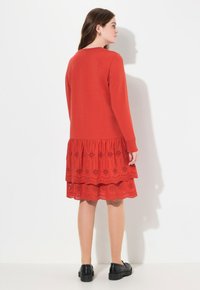 Rotes Kleid mit langen Ärmeln und einem gestuften Spitzenrüschensaum. Die Spitze zeigt durchbrochene florale Muster. Tragen mit schwarzen Schuhen.