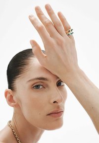 Tres anillos apilados en los dedos del modelo, con acentos en oro y verde. La mano se muestra contra un fondo neutro.