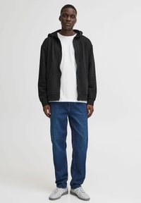Black zip-up hoodie over a white t-shirt, paired with blue denim jeans and gray sneakers. The hoodie has a relaxed fit and pockets.