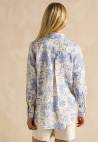 Kobieta z długimi blond włosami, ubrana w koszulę z długim rękawem w niebiesko-białe wzory oraz jasne szorty, stojąca przy beżowej ścianie.