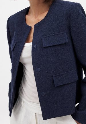 Femme portant une veste texturée bleu marine boutonnée avec des poches à rabat sur un débardeur blanc et un pantalon clair, sur un fond uni.