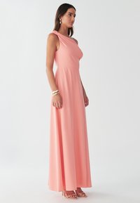 Sieviete tērpusies bezroku korāļrozā viena pleca maksi kleitā ar bēšām augstpapēžu sandālēm un zelta aprocēm uz plaukstas.