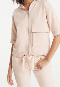Felpa con zip di colore rosa chiaro, maniche corte, tasca frontale, vita con cordoncino e texture liscia. Presenta dettagli metallici sulla zip.