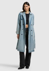 Nő, aki hosszú, világoskék farmer ballonkabátot visel fekete croptoppal, fekete, magasderekú, bő szárú farmernadrággal és fekete hegyes orrú csizmával.