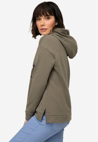 Olivgrön hoodie med dragsko, mjuk textur, sidslitsar vid fållen, bärs med ljusblå jeans, sedd från en lätt sidovinkel.