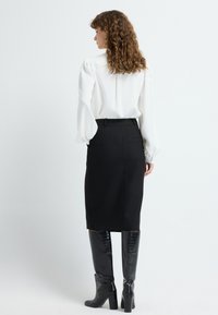 Blouse blanche avec des manches ballons, associée à une jupe noire ajustée au genou et des bottes noires brillantes jusqu'au genou.