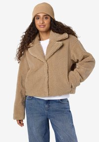 Dzeltenbrūns mākslīgās kažokādas žakete ar apkakli, saīsinātu dizainu un platajiem piedurknēm, kombinēta ar pieguļošu ribotu cepuri. Valkāts pār baltu kreklu.