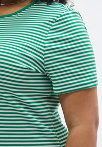 Žalia baltais dryžiais marginta marškinėliai su apvalia iškirpte, trumpomis rankovėmis ir minkšta medvilnės medžiaga. Dryžiai tolygiai išdėstyti.