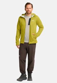 Groene lange mouwen jack met capuchon, gecombineerd met een grijs T-shirt en bruine broek. De outfit heeft ritsen en gestructureerde materialen.
