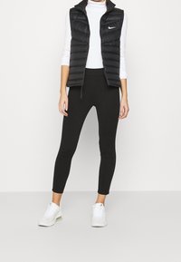 Svart puffärmad väst med hög krage och dragkedja, vit långärmad topp, svarta leggings och vita sneakers med ljusa accenter.