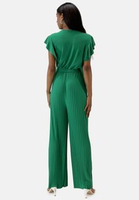 Mono verde con pliegues y mangas de volantes, cintura ceñida y diseño de pierna ancha. Tejido suave con un brillo sutil, combinado con zapatos de tacón.
