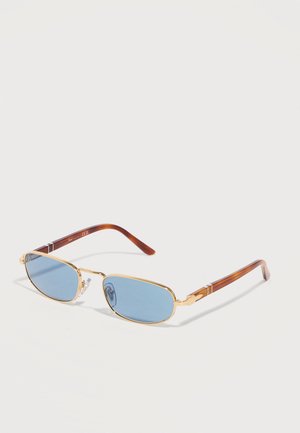 Pravokutne naočale s plavim nijansama, zlatnim metalnim okvirom i smeđim krakovima s uzorkom kornjačevine koje leže na svijetloj površini.