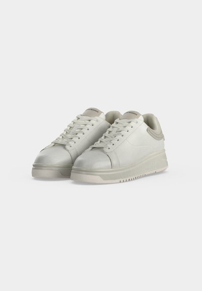 Ζευγάρι λευκά δερμάτινα sneakers χαμηλού προφίλ με κορδόνια και παχιά λαστιχένια σόλα σε απλό ανοιχτό γκρι φόντο.
