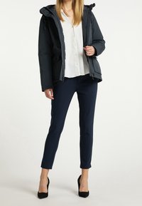 Svart isolerad jacka med huva, knappad framtill och kviltad textur, kombinerad med en vit blus och mörkblå tajta byxor, kompletterad med svarta klackskor.