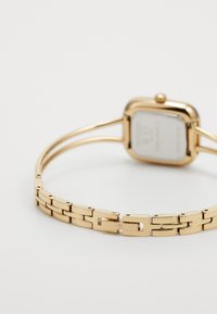 Gold metal watch with a rectangular case, linked bracelet, and smooth texture. The back features minimal text and a seamless clasp design.