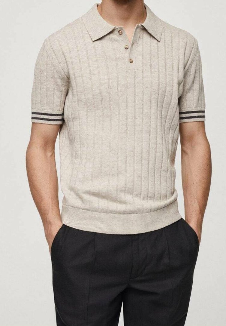 Homme portant un polo en maille beige à manches courtes avec des côtes verticales et des poignets rayés noirs, associé à un pantalon noir et les mains dans les poches.