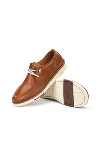 Chaussures décontractées en cuir marron à enfiler avec semelles en caoutchouc blanches texturées, une chaussure debout et l’autre montrant l’empreinte de la semelle.