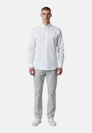 Giovane uomo in piedi di fronte, che indossa una camicia bianca con bottoni, pantaloni grigio chiaro e scarpe da ginnastica bianche su uno sfondo semplice.