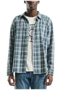 Pārbaudes raksta pogām piesprausta krekls zilo un zaļu toņu kombinācijā, ar brīvu piegriezumu, krūšu kabatu un vieglu audumu, uz kura ir uzlikta grafiskā t-kreklu.