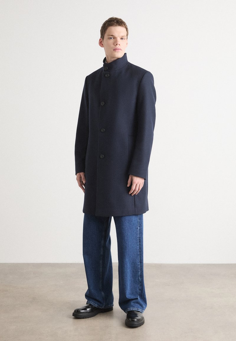 Cappotto di lana navy con colletto alto, dotato di bottoni frontali, tasche laterali e taglio dritto. Abbinato a jeans denim a gamba larga e scarpe nere.