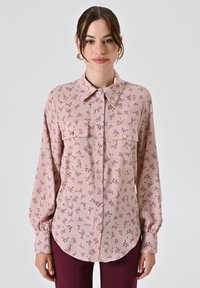 Blusa rosa chiaro con motivo floreale, due tasche sul petto, chiusura a bottoni e maniche lunghe. Il materiale appare liscio e leggermente lucido.