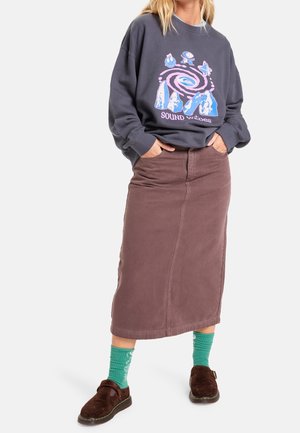 Kvinde iført mørk grafisk sweatshirt, brun midi nederdel, grønne mønstrede sokker og mørkebrune træsko, stående med hænderne i lommerne.