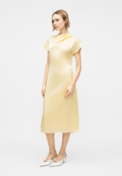 Γυναίκα με παλ ροζ σατέν midi φόρεμα με κοντά μανίκια και άσπρα μυτερά τακούνια με λουράκια στον αστράγαλο, στέκεται μπροστά σε απλό λευκό φόντο.