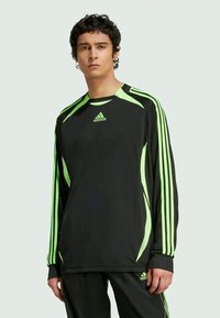Jaunas vyras vilkintis juodas ir neoninėmis žalia sportinėmis Adidas ilgomis rankovėmis palaidine bei suderintomis kelnėmis stovi prieš paprastą šviesų foną.