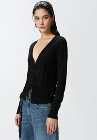 Cardigan nero con scollatura a V profonda, cinque bottoni e polsini a costine. Indossato sopra jeans azzurri chiari, evidenziando un design aderente e una texture liscia.