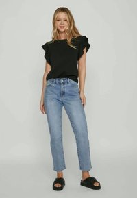 Woman wearing a black ruffled sleeve top, light blue straight-leg jeans, and black slip-on sandals, standing against a plain background.