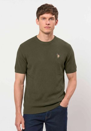 Homme aux cheveux courts et bruns portant un pull en maille à manches courtes vert olive avec un petit logo brodé sur la poitrine et un jean bleu foncé.