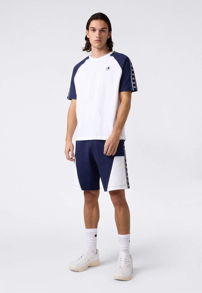 T-shirt en coton texturé blanc avec des manches bleu marine ; associé à un short bleu marine et blanc avec un grand panneau latéral et des accents de logo.