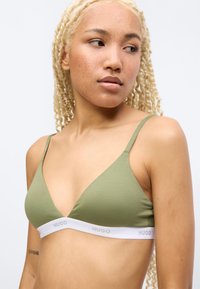 Νεαρό άτομο με ανοιχτόχρωμο δέρμα και πλεξούδες ξανθά μαλλιά φοράει πράσινο τρίγωνο σουτιέν με λευκή ελαστική ταινία που φέρει την επιγραφή "HUGO," κοιτάζοντας πλάγια.