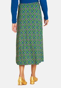 Grüne, floral gemusterte Midirock mit blauen und rosa Akzenten. Das Material hat eine strukturierte Oberfläche. Tragen mit goldenen Blockabsätzen.