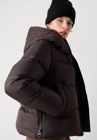 Une personne portant une doudoune marron foncé avec un col haut et un bonnet noir, regardant par-dessus son épaule devant un fond uni.