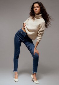 Krémový pletený sveter s vysokým golierom, kombinovaný s tmavomodrými obtiahnutými džínami s rozstrapkanými lemy a béžovými vysokými podpätkami.