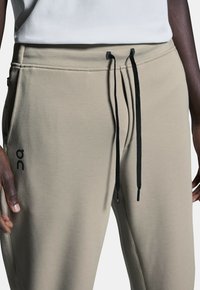 Beige sweatpants met een gladde textuur, voorzien van een zwarte trekkoord en een logo-accent aan de linkerkant. Aangesloten pasvorm met zijzakken.