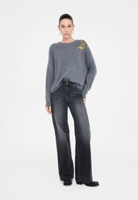 Maglione grigio lavorato a maglia con una finitura testurizzata e un dettaglio di perline su una spalla, abbinato a jeans a gamba larga grigio scuro a vita alta.