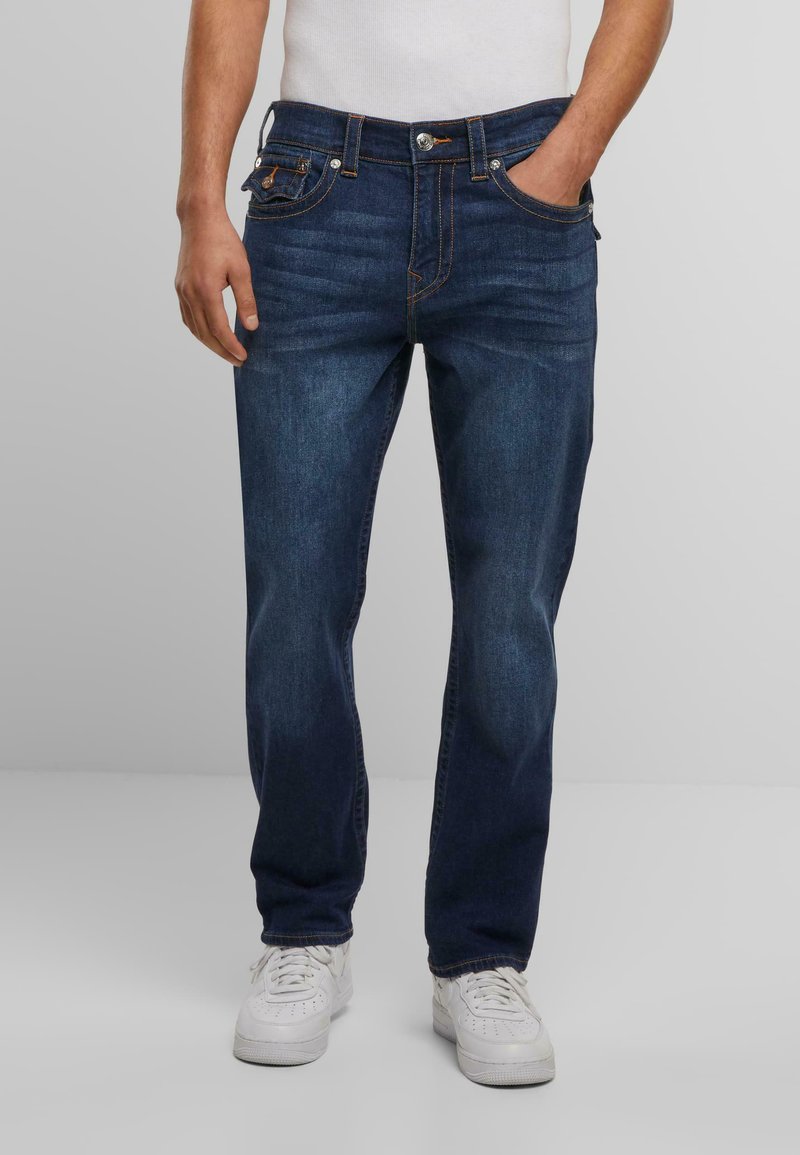 Donkerblauwe denim jeans met een rechte pasvorm, voorzien van vijf zakken, oranje stiksels en een knoop/ritsluiting. Gemaakt met witte sneakers.