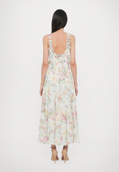 Een bloemenmaxi-jurk met een lage rug, voorzien van ruffles op de schouderbanden, pastelkleuren en een geplooide rok. Het materiaal lijkt licht en zacht.